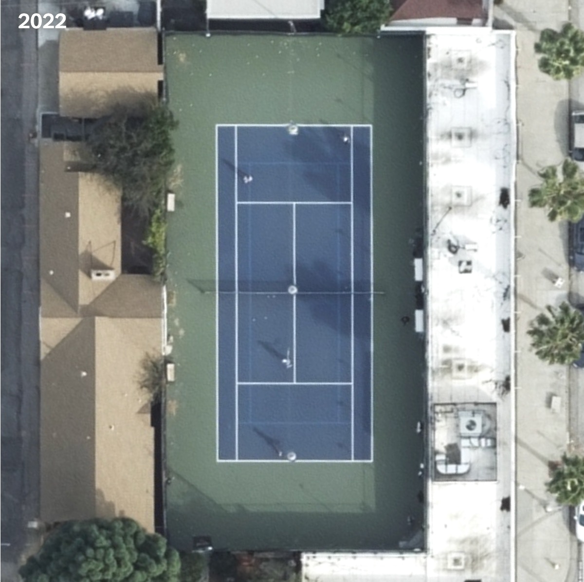 Aerial view of a tennis court surrounded by buildings, with players visible on the court. The scene is from 2022.